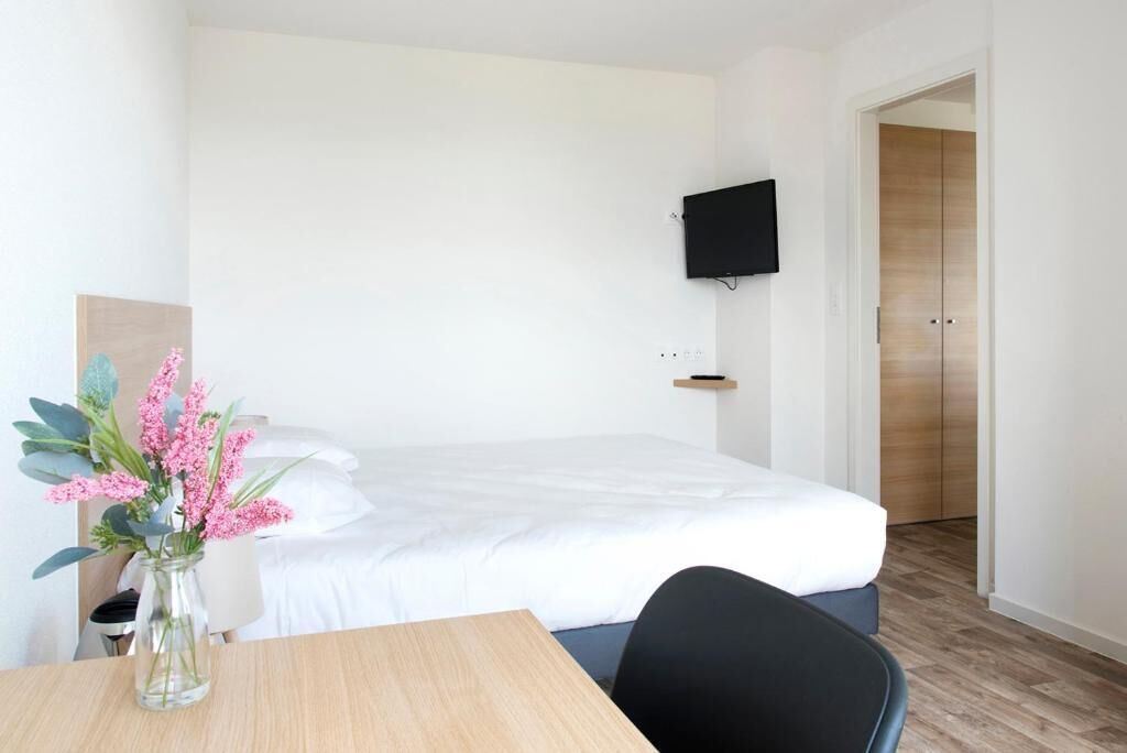 Quarto de hotel com cama, televisão na parede, piso de madeira e uma pequena mesa com um vaso de flores.