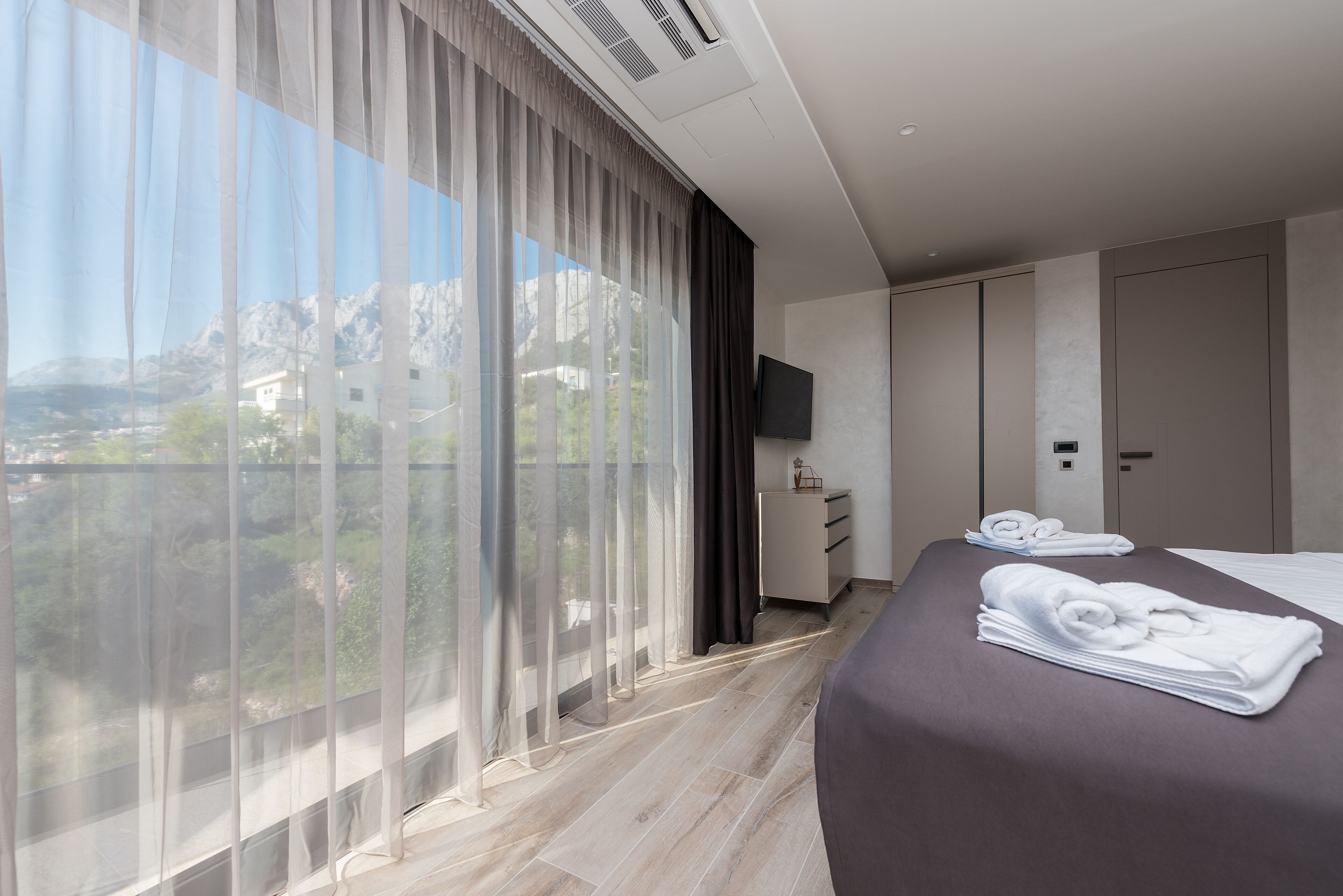 A modern hotel room with a large window, a bed with neatly folded towels, a flat-screen TV, and a wardrobe.