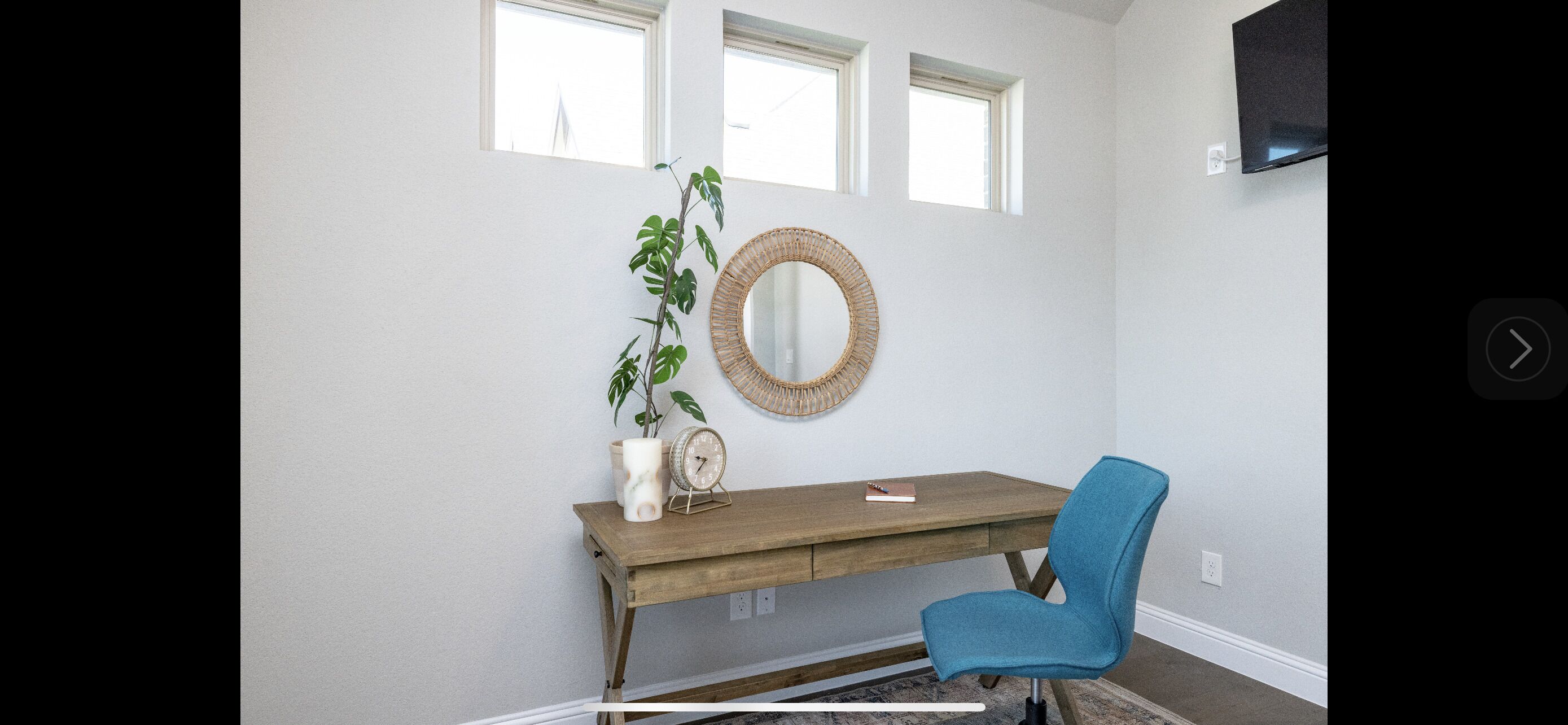 A room with a wooden desk, a blue chair, a potted plant, a round mirror, and a television mounted on the wall.