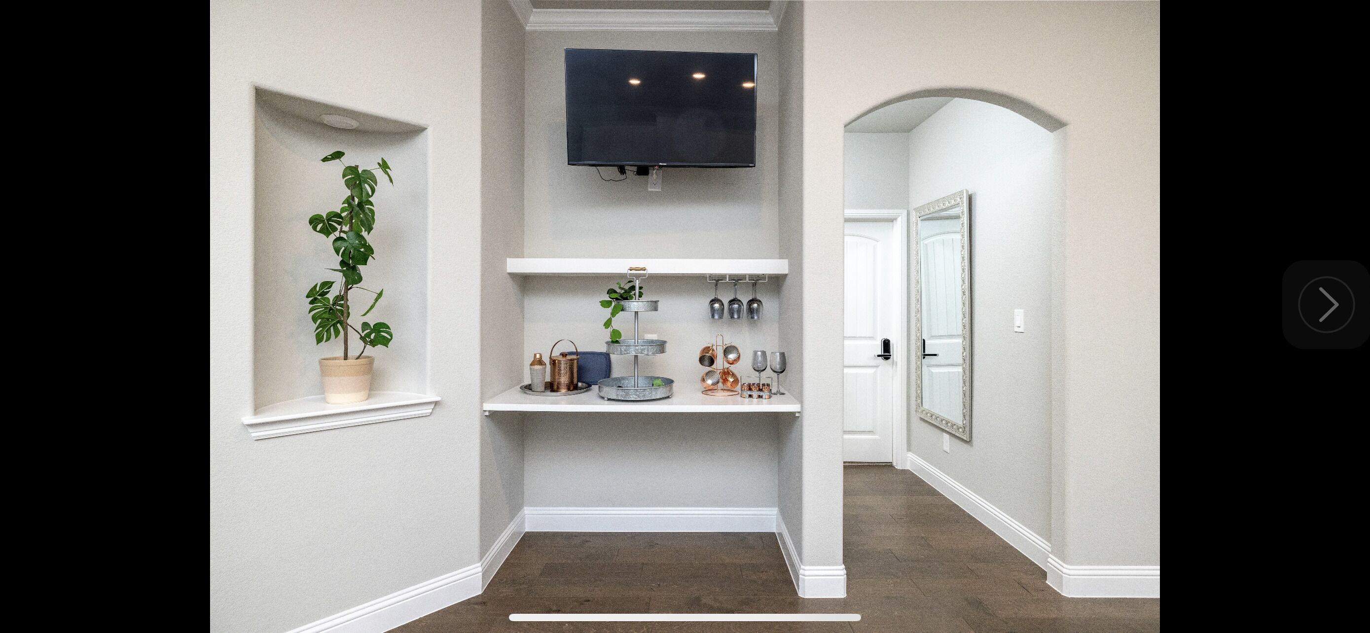 Un couloir avec un téléviseur fixé au mur, une plante sur le rebord d’une fenêtre et une étagère ornée d’objets décoratifs.