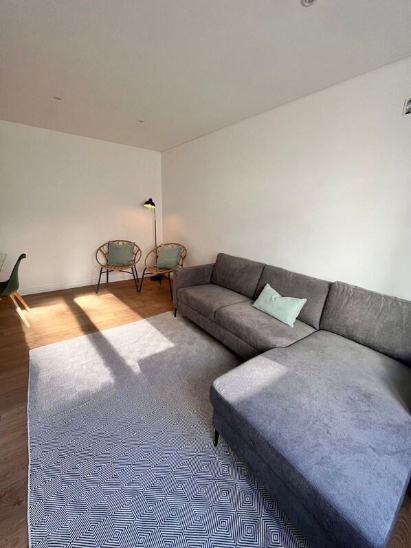 Ein modernes Wohnzimmer mit einem grauen Ecksofa, zwei Korbstühlen, einem gemusterten Teppich und einer Stehlampe.