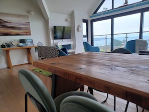 Un salon moderne avec une table à manger en bois, des sièges confortables, un téléviseur à écran plat et une vue sur la mer grâce à de grandes fenêtres.
