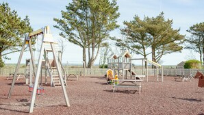 A playground with wooden structures, including a swing set and climbing frames, surrounded by trees and a fenced area.