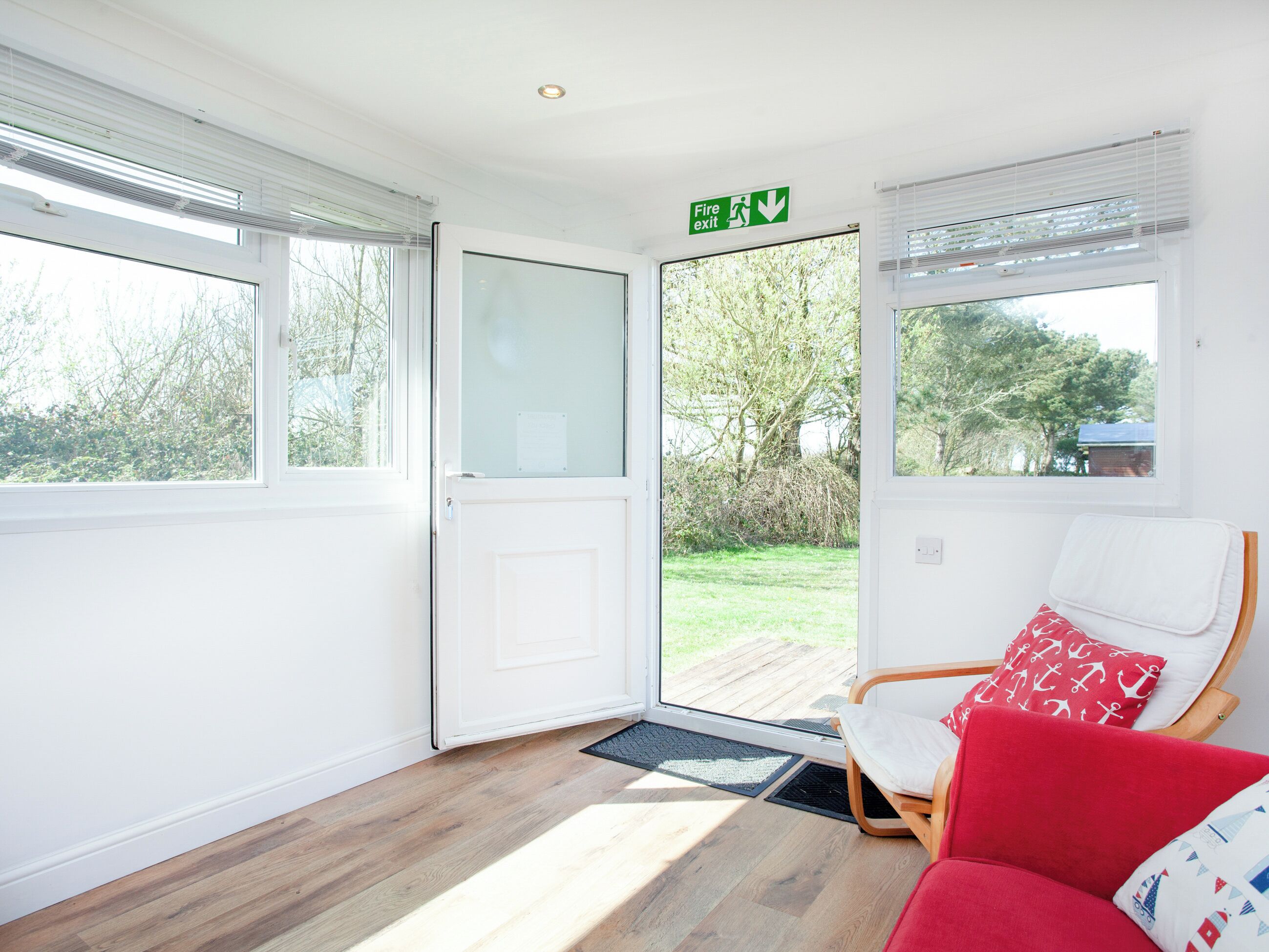 A room with a wooden floor, a white door, a red chair, and a view of greenery outside.