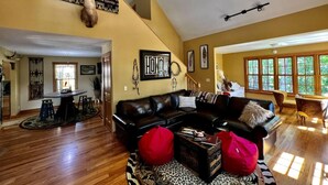 A spacious living room with a black leather sofa, a red bean bag, and a wooden coffee table. There is a ceiling fan, a wall-mounted deer head, and a large window providing natural light. - Brown County At It's Best Beautiful Cozy meticulous clean LG Game Room arcade! (Nashville)