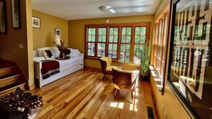 A spacious living room with a wooden floor, a white sofa, a dining table with chairs, and large windows offering a view of greenery outside. - Brown County At It's Best Beautiful Cozy meticulous clean LG Game Room arcade! (Nashville)