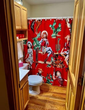 Une salle de bain avec un rideau de douche rouge orné d’un motif représentant un cactus et une femme, des meubles en bois, des toilettes blanches et un plancher en bois.