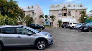 A parking lot with several cars, a building with a light-colored facade and multiple balconies, and palm trees in the background. - Magnificent seaside studio (Gosier)