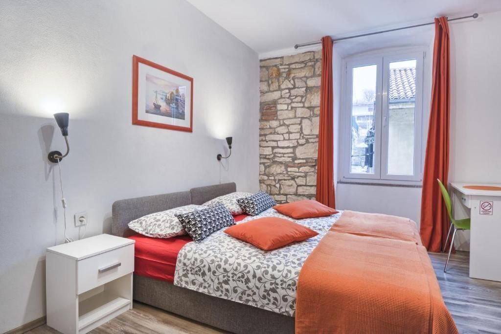 Ein Schlafzimmer mit Bett, Nachttisch, Schreibtisch, einer Steinsäule und einem Fenster mit Vorhängen.