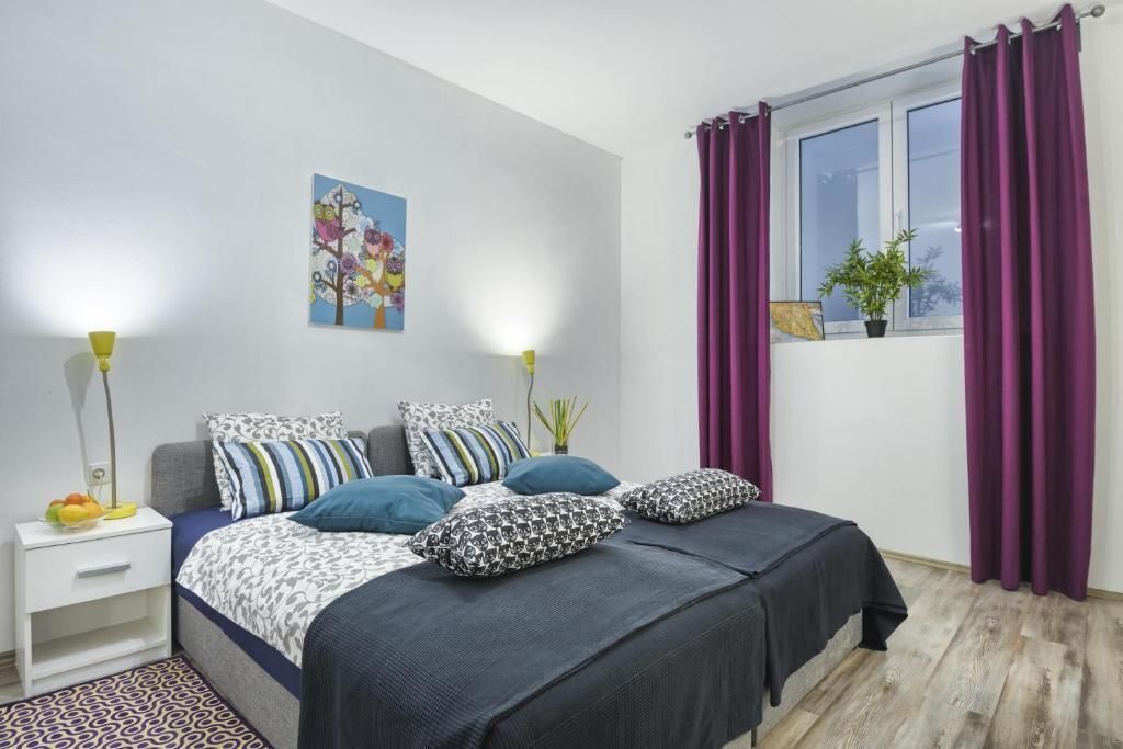 A bedroom with two beds, a bedside table with a lamp, a window with purple curtains, and a wall-mounted plant.