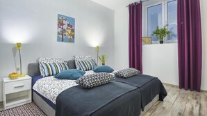 Une chambre avec deux lits, une table de chevet avec une lampe, une fenêtre avec des rideaux violets et une plante fixée au mur.