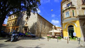 Une rue bordée d’un bâtiment en pierre, de voitures garées et d’un bâtiment jaune doté d’une arche.