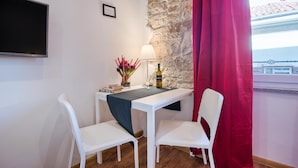 Una habitación pequeña con una mesa blanca, dos sillas blancas, un televisor montado en la pared, una pared de piedra y una cortina roja.