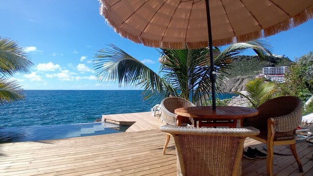 Una terraza de madera con mobiliario de mimbre y una sombrilla, con vistas a un mar azul cristalino y una costa rocosa.