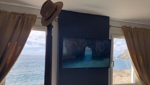 Una habitación con un ventanal que ofrece vistas al mar, un televisor de pantalla plana en la pared y un sombrero decorativo colgado en el marco de la ventana.