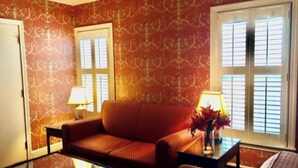 Ein Zimmer mit einer Couch, einem Glastisch und zwei Lampen. Es gibt zwei Fenster mit Fensterläden.