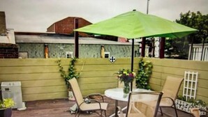 Ein Außenbereich mit Terrasse, einem grünen Sonnenschirm, zwei Stühlen, einem kleinen runden Tisch und Topfpflanzen.