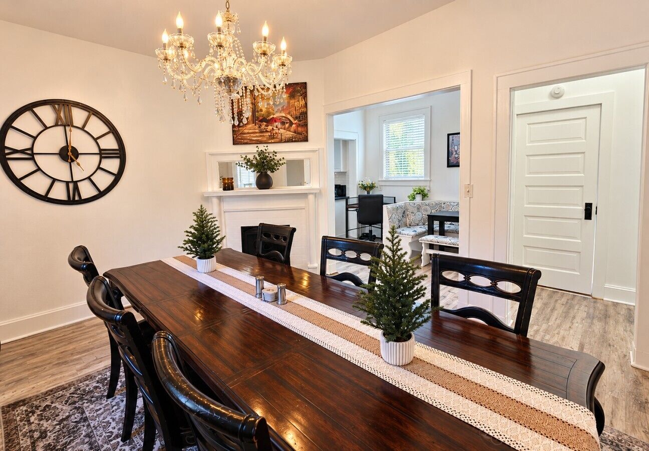 A dining room with a long wooden table, black chairs, a large wall clock, a chandelier, and a Christmas tree.