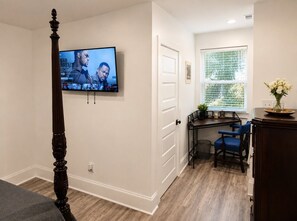 A room with a television mounted on the wall, a dining area with chairs, a window with blinds, and a door. - Historic Victorian Restoration (Savannah)