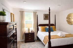 A bedroom with a four-poster bed, a nightstand with a lamp, a dresser with drawers, and a window with curtains.