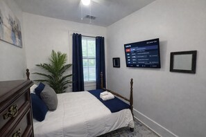 Ein Schlafzimmer mit einem Bett, einer Kommode, einer Topfpflanze, einem Fenster mit Vorhängen und einem an der Wand montierten Fernseher.