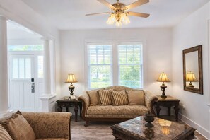 A room with a ceiling fan, two windows with blinds, a sofa, two side tables with lamps, and a door.
