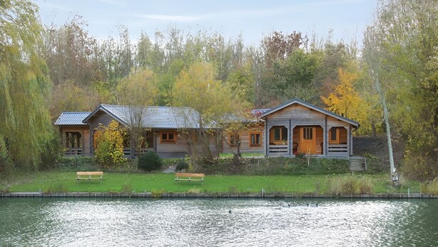 Two wooden cabins by a lake, with benches in front and surrounded by trees. - Chalet Copperwing on small-scale nature campsite 't Ol Gat. (Zoutkamp)