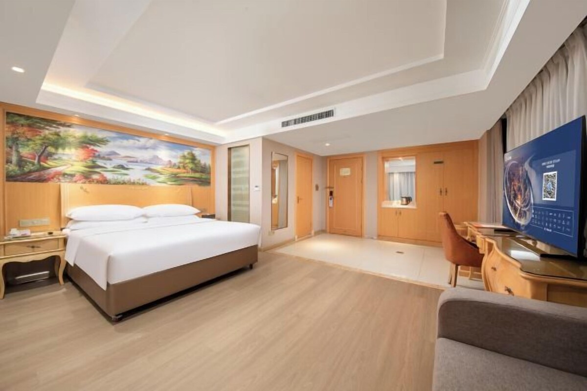 Quarto de hotel com uma cama, escrivaninha, televisão e um quadro na parede.