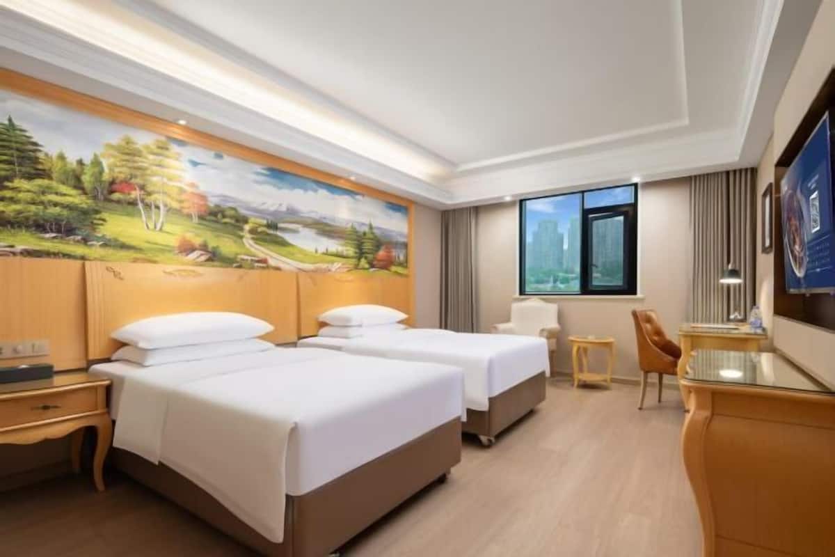 A hotel room with two beds, a desk, a chair, a painting on the wall, and a window with a city view.