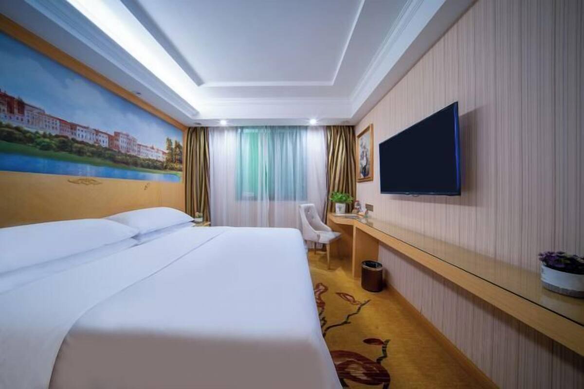 A hotel room with a bed, a desk, a chair, a television, and a painting on the wall.