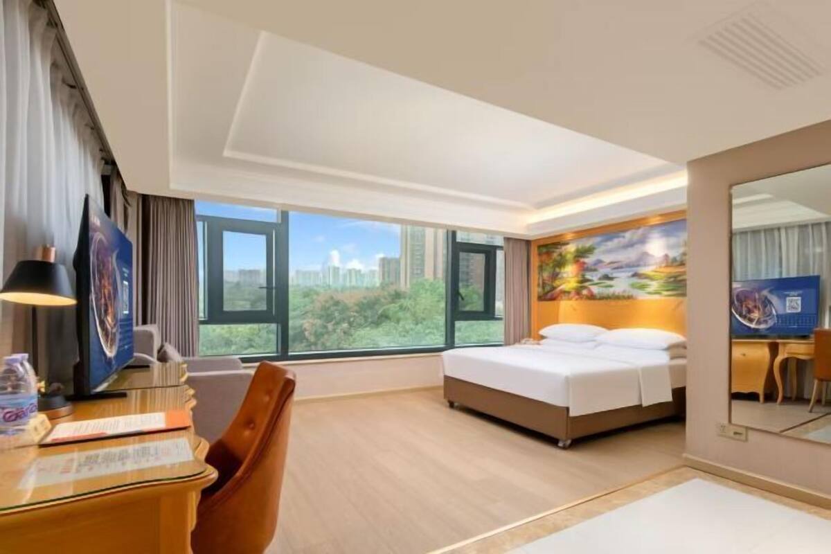 A hotel room with a bed, desk, chair, large windows, and a painting on the wall.