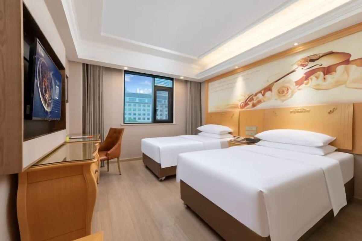 A hotel room with two beds, a desk, a chair, a large window, and a wall-mounted TV.