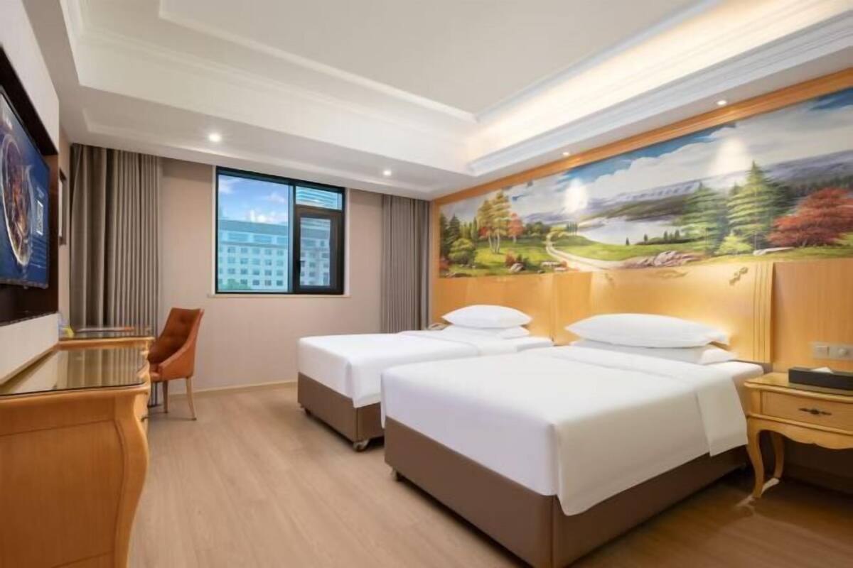 A hotel room with two beds, a desk, a chair, a painting on the wall, and a window with a view of a building.