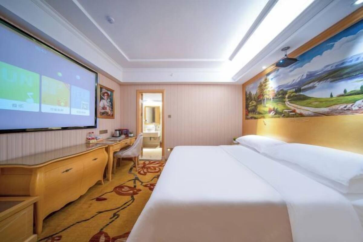 A hotel room with a bed, desk, chair, and a large screen displaying content.