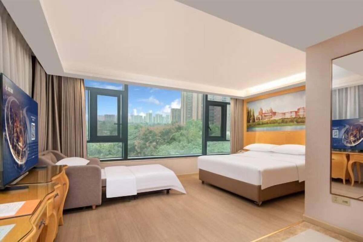 A hotel room with a bed, a desk, a chair, and a large window offering a city view.
