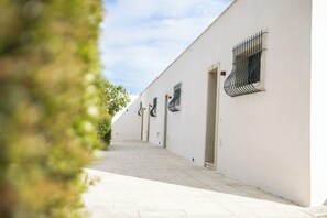 Un étroit sentier mène à un bâtiment aux murs blancs et aux nombreuses fenêtres ornées de barreaux en métal noir.