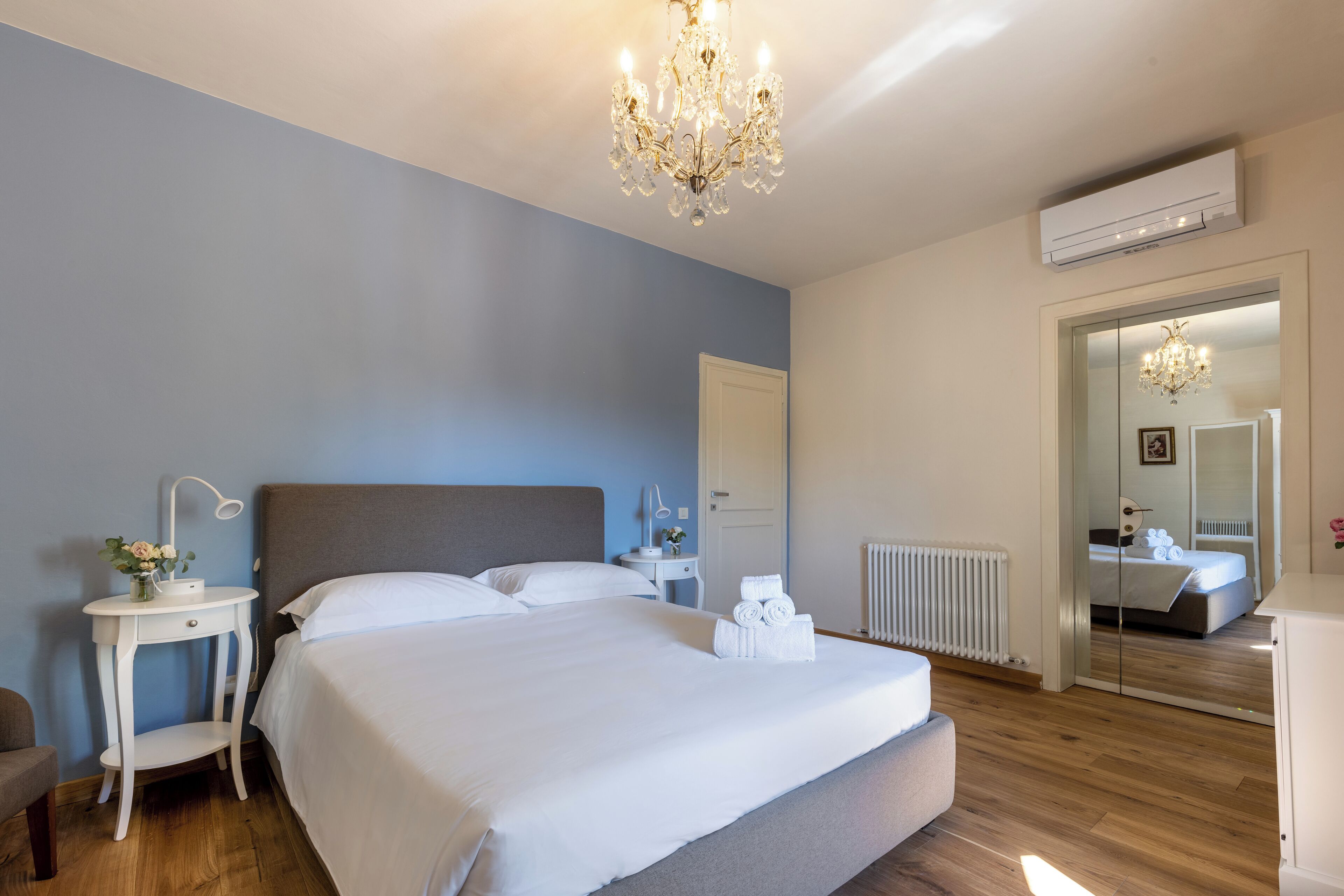 A hotel room with a large bed, bedside tables, a chandelier, an air conditioning unit, and a mirror reflecting the room.