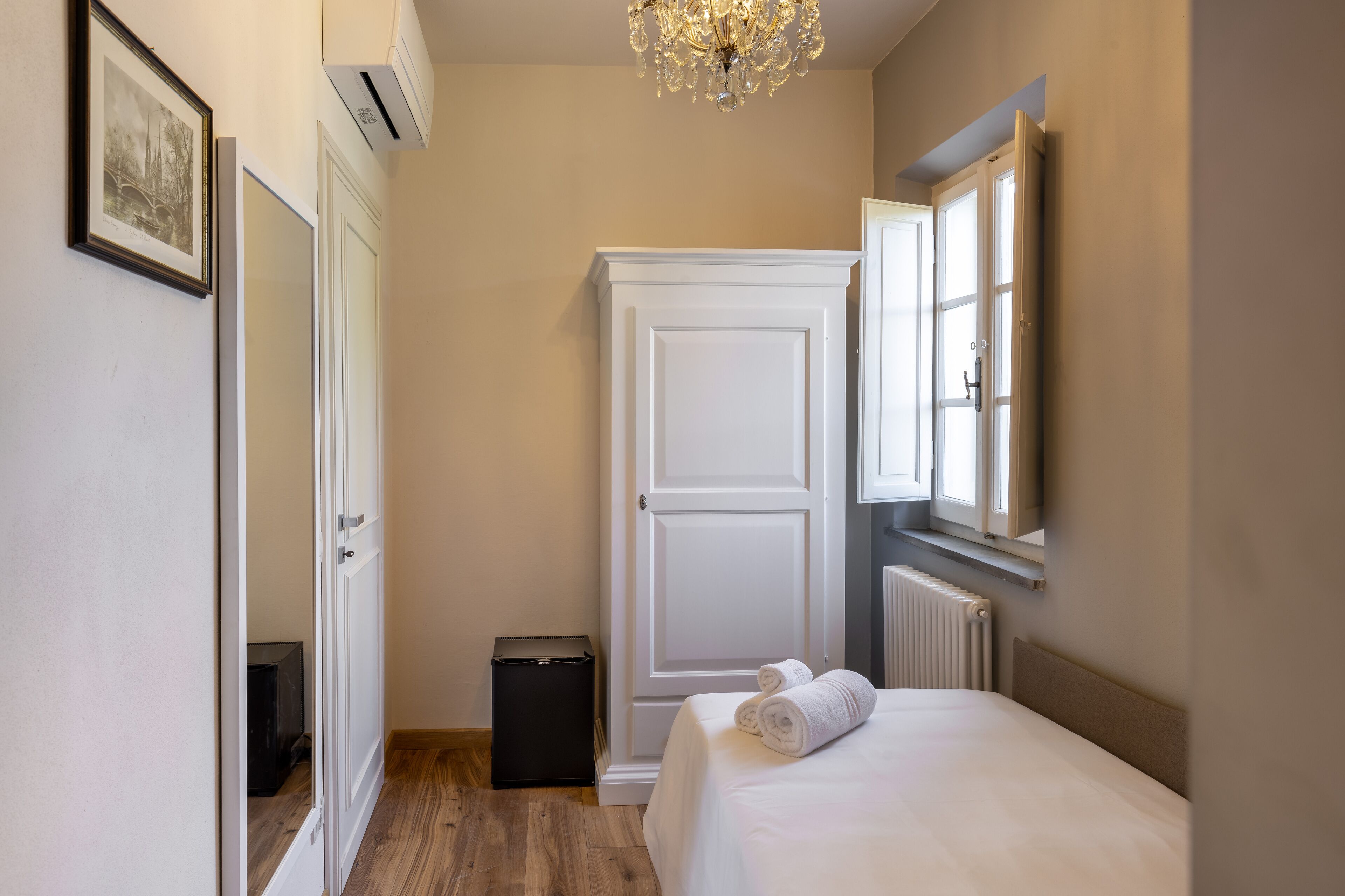 A bedroom with a bed, a radiator, a white wardrobe, a small trash bin, and a framed picture on the wall.