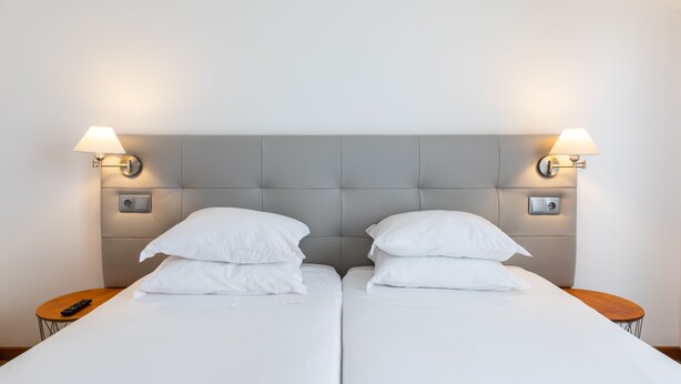 A double bed with a grey upholstered headboard, two bedside tables with lamps, and a remote control on one of the tables. - Arcangel Hotel (Lagoa)