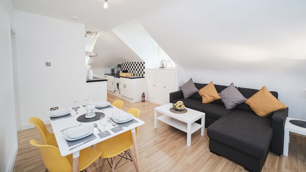 A modern apartment with a dining area, a black sofa with cushions, and a kitchen in the background.