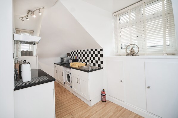 A kitchen with white cabinets, a black countertop, a checkered backsplash, and a window with blinds. - Flat 5 - 61 Old Dover Road (Canterbury)