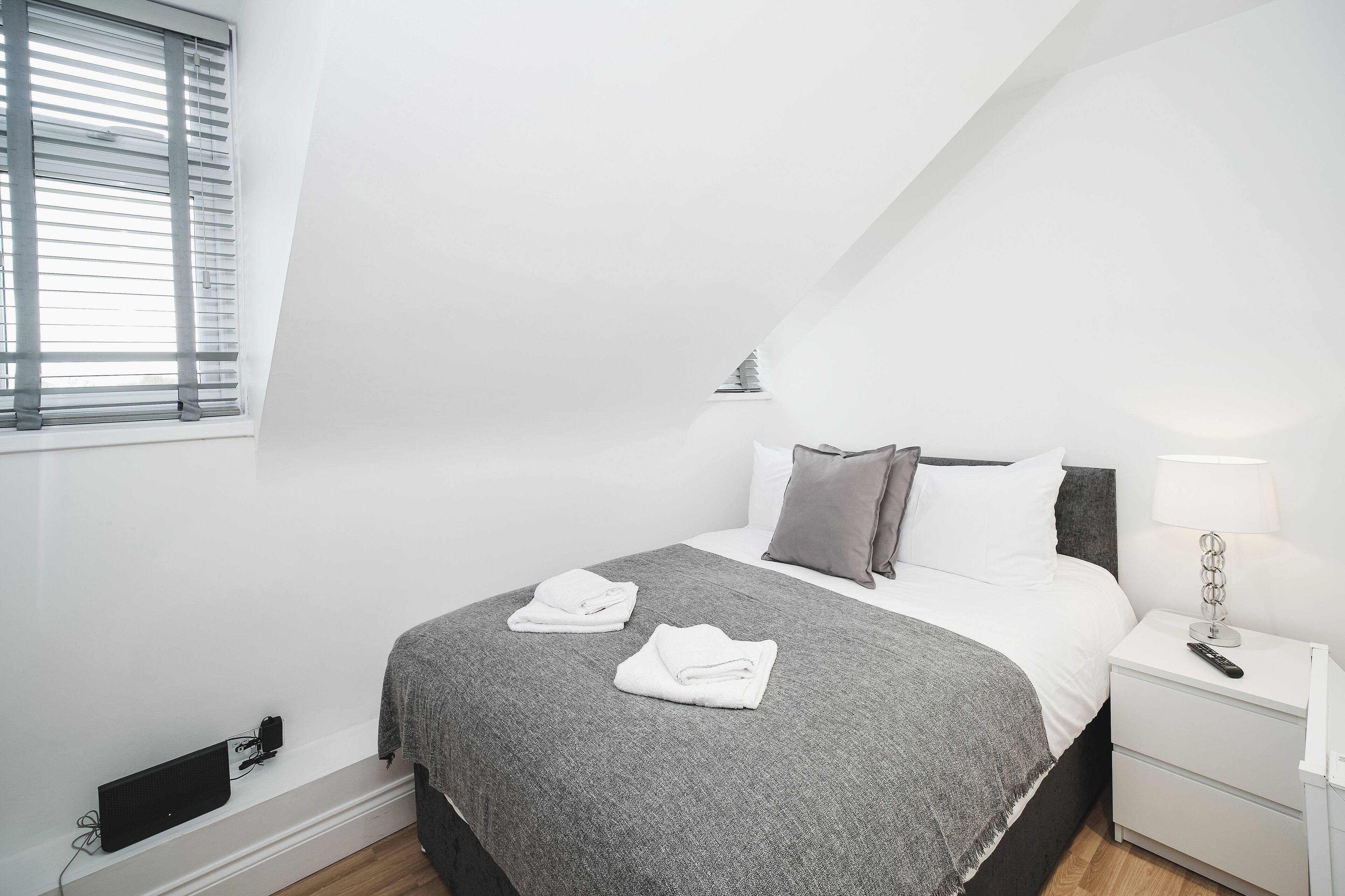 A neatly made bed with a grey blanket and two folded white towels, a bedside lamp, and a power outlet on the nightstand.