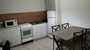 Et lille køkken med hvide skabe, en mikrobølgeovn, komfur og køleskab. Der er et spisebord med stole og en hængende lampeskærm.