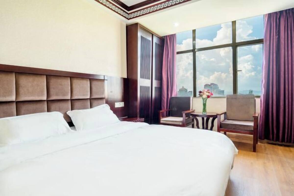 Une chambre d’hôtel avec un grand lit, une tête de lit en bois, une fenêtre avec des rideaux, une petite table avec un vase et une chaise.