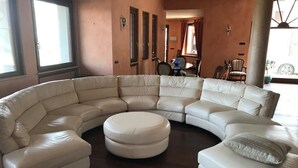 A large, L-shaped sectional sofa with a round ottoman in a room with wooden flooring and large windows. - Remarkable 6 Bedrooms Villapiemonte With Land (Cerrione)