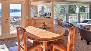 Uma sala de jantar com uma mesa de madeira redonda e cadeiras, um espelho grande e vista para um deck e para a água lá fora.
