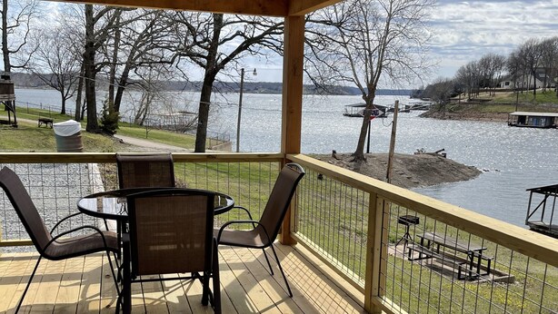 A covered deck with a table and chairs, overlooking a lake with a dock and picnic tables. - Cozy lake front Cabin that's perfect for two! (Grove)
