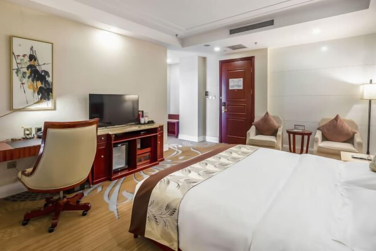 A hotel room with a large bed, a desk with a computer, a TV, and two armchairs.