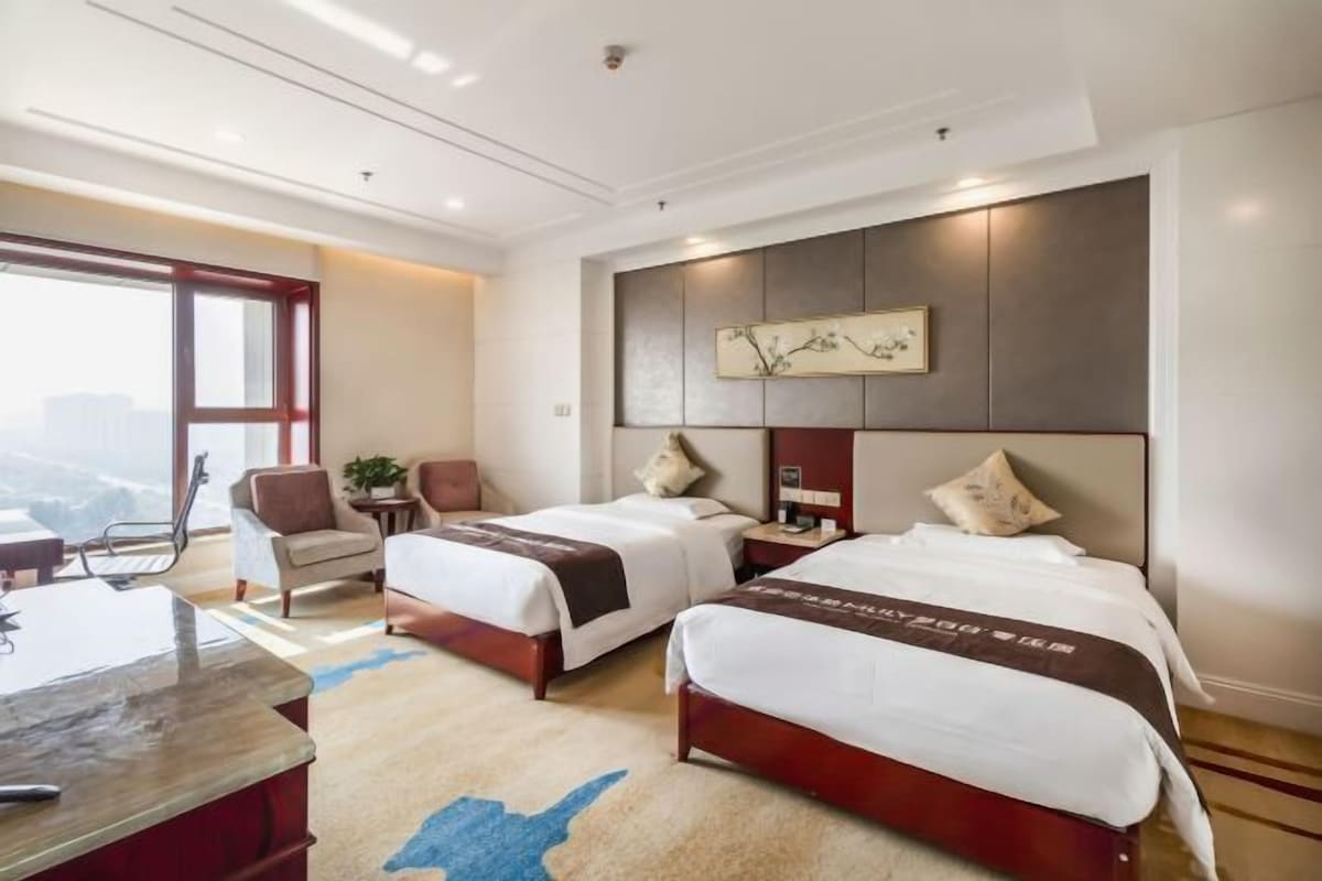 A hotel room with two beds, a desk, and a large window.