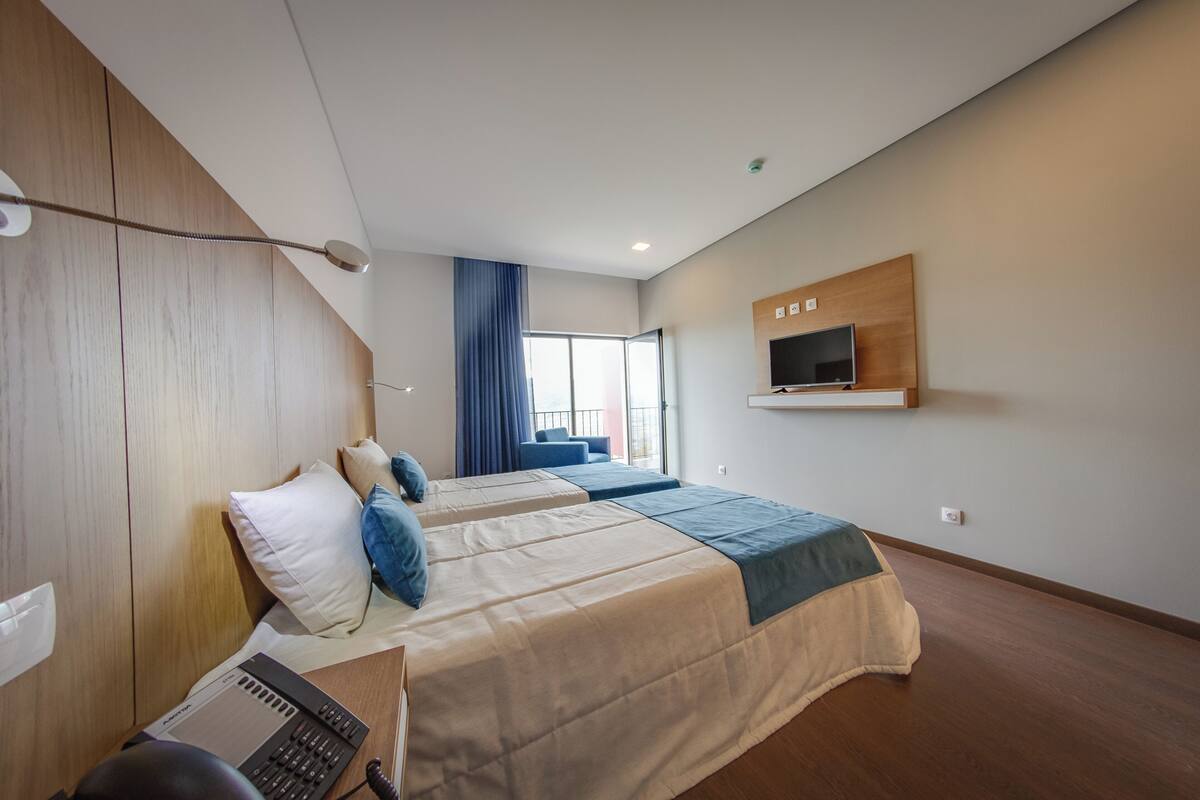 A modern hotel room with two beds, a desk with a computer, a TV mounted on the wall, and a large window with curtains.